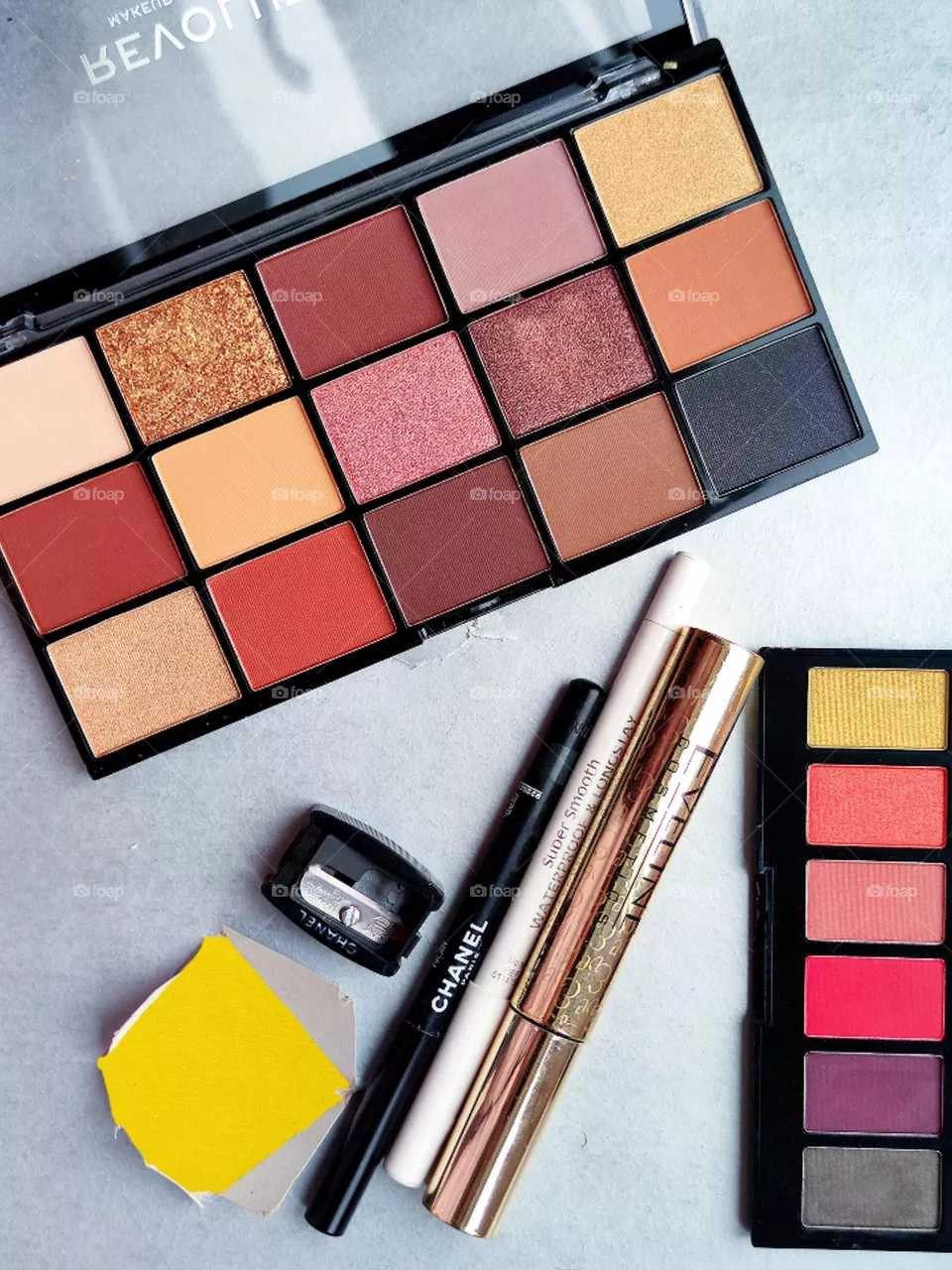 Cosmetics for the eyes. On a gray background are: two multi-colored eyeshadow palettes, a black pencil, mascara, a corrective white pencil and a sharpener. In the corner of the background is a hole with a yellow background. Contrast