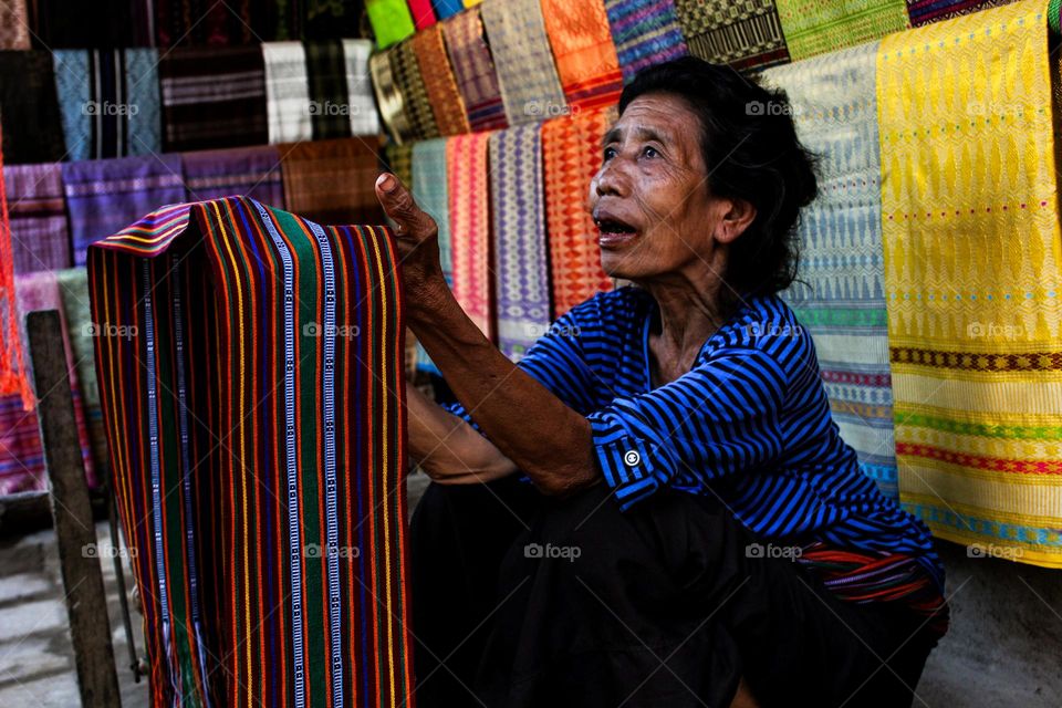 Portrait of an old woman sitting holding lombok woven cloth for sale as traditional souvenirs from Sade village, traditional cloth display in the background, Lombok, Indonesia.