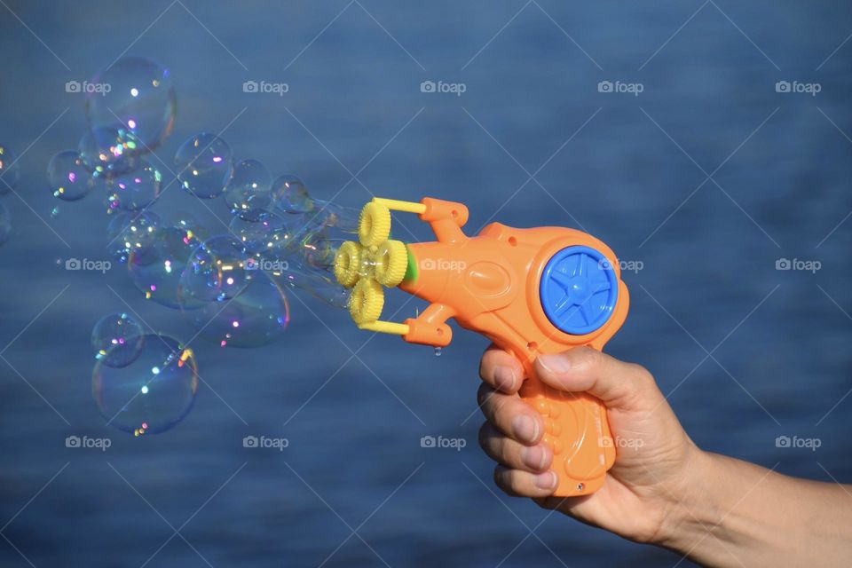 Close-up of hand holding a soap bubbles gun in orange and blue colors against blue background. 