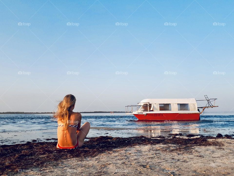 Faceless girl in swimming suit sits alone at the beach in front of boat