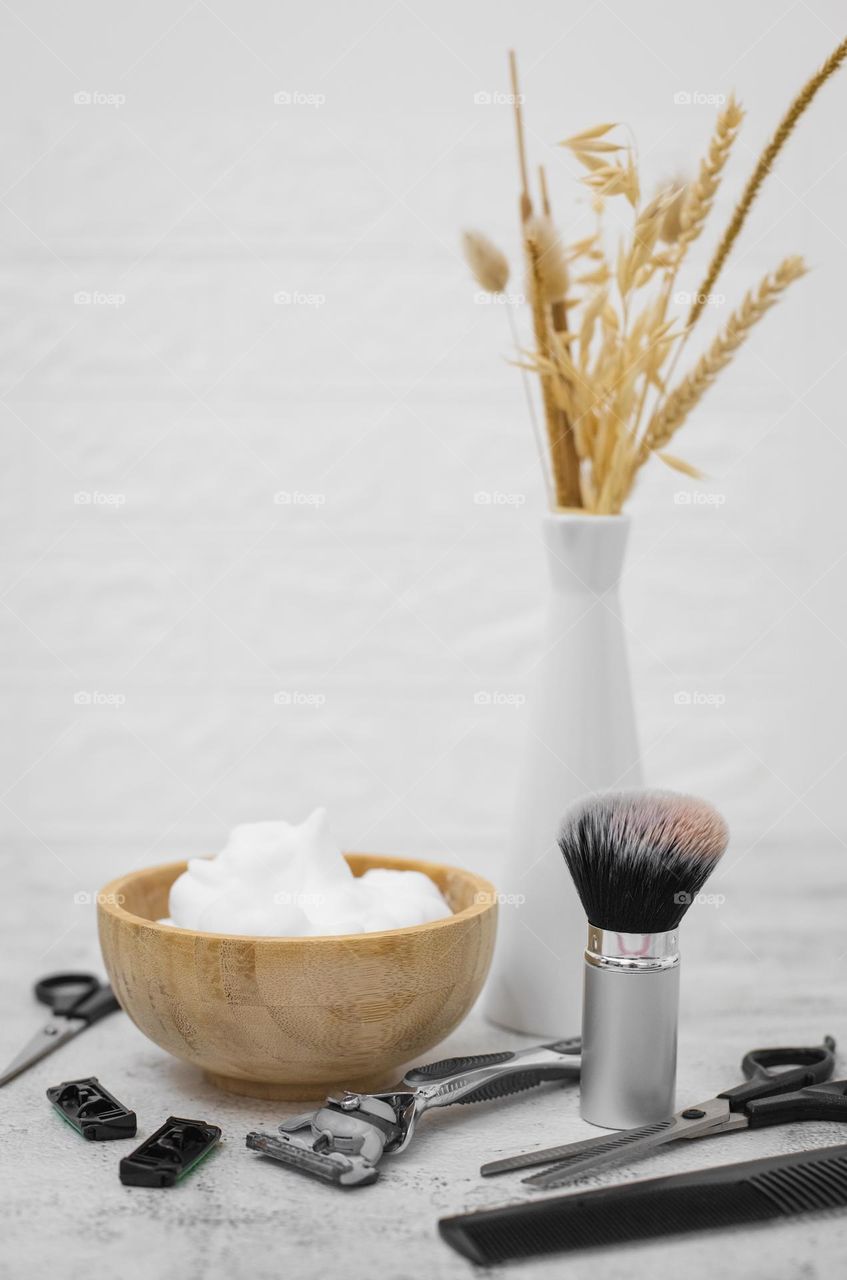 Wooden bowl with shaving foam, fluffy shaving brush made of natural fleece,forehead vase with dried flowers, razor,nozzles,scissors and comb on a light stone background with a white blurred brick wall,side view. Concept barbershop man,shaving man,bea