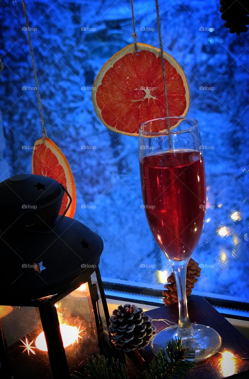 A glass of red wine stands on the windowsill. Next to it is a lantern with a luminous candle and pine cones. Dried oranges and cones hang on the window. Outside the window trees in the snow