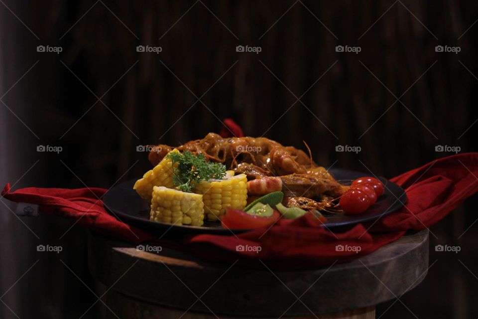 Asian food balado fried prawns with corn chunks, lemon slices and tomatoes served on black plate with red cloth bottom and wooden coaster. Asian style street seafood.