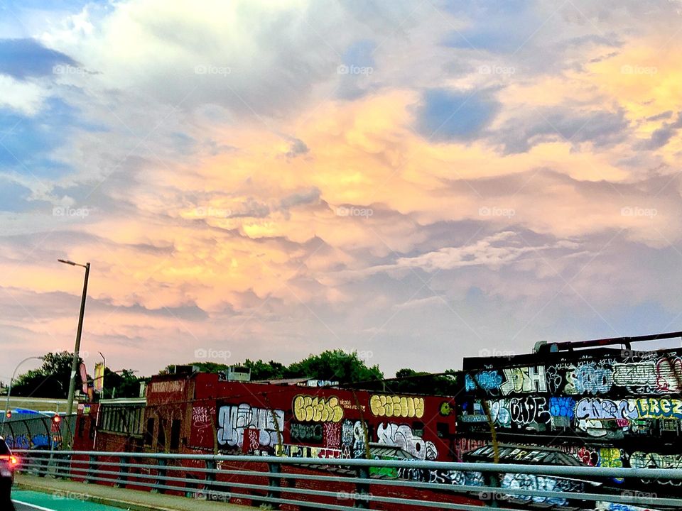 Graffiti, dumpsters & sheds next to a road in LIC, Queens can take on a streamlined beautiful appearance when the rays of the setting sun meet the manifold cumulus clouds above and reflect them back down to us. 2021. Hypnotic Productions