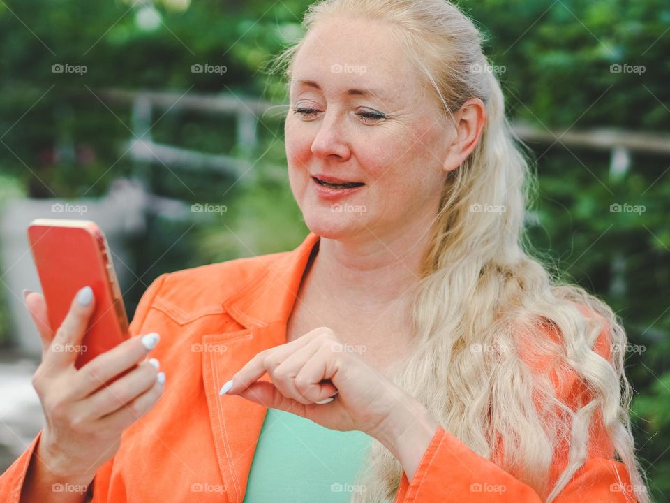 Portrait of a beautiful young blonde woman in an orange jacket with a mobile phone in her hands sitting at a street cafe table on a summer day, close-up side view.Concept using a smartphone.