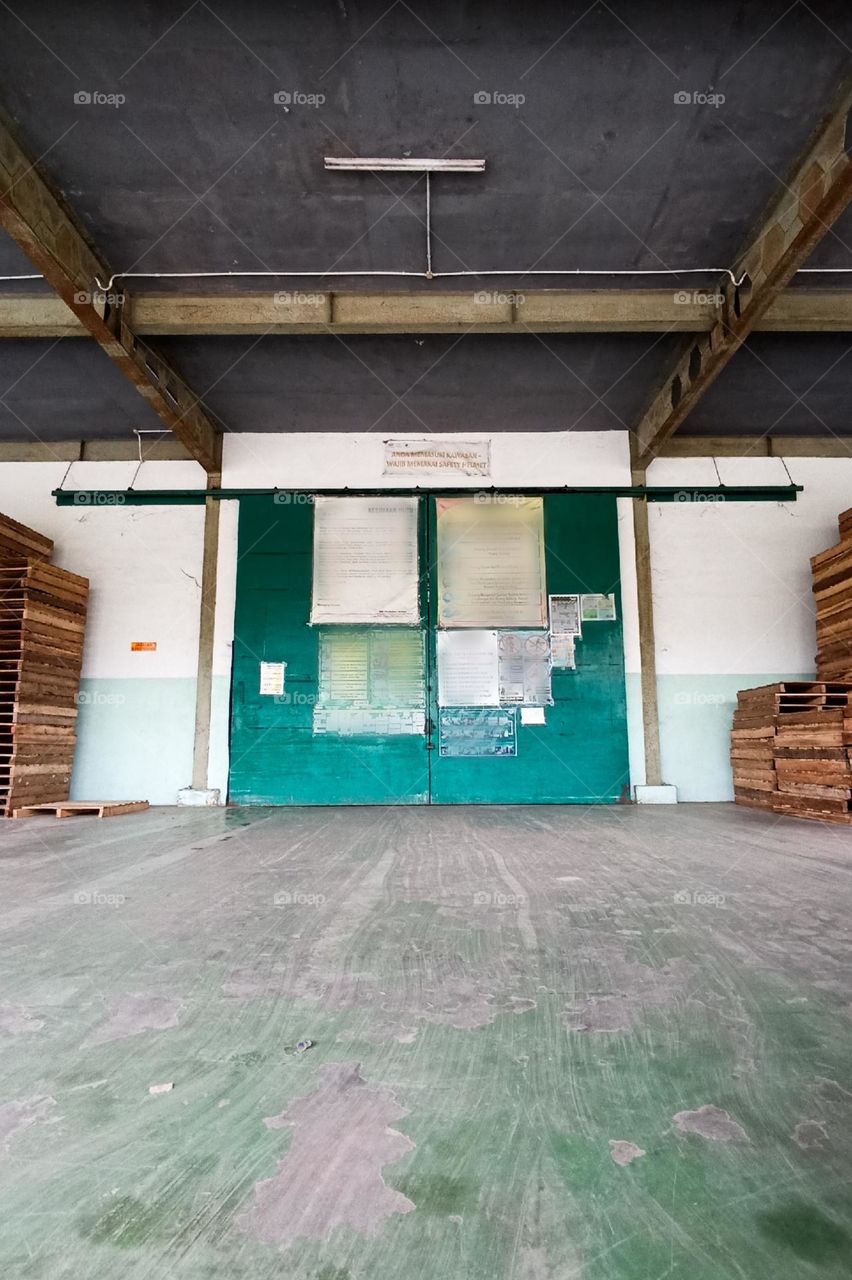 warehouse or storage area with a large green door on the back wall, concrete floor, and several wooden pallets arranged on the sides