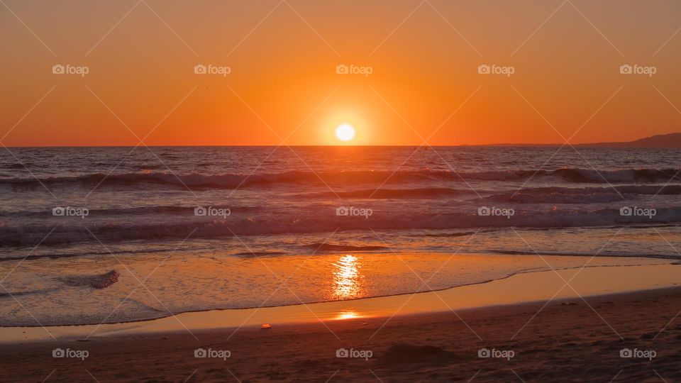 Sun setting into water horizon at Santa Monica Beach in Los Angeles, CA, USA. Warm natural light of the sunset twilight reflects beautifully onto the calm water with small waves. Romantic nature photo.