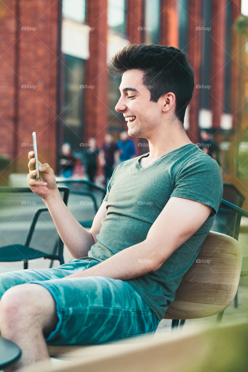 Young man having fun with smartphone, reading funny texts, sitting in center of town