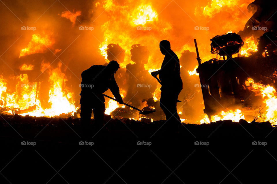 Bright flames illuminate the night, while two fire fighters work to clear debris from the highway following a tanker rollover. 