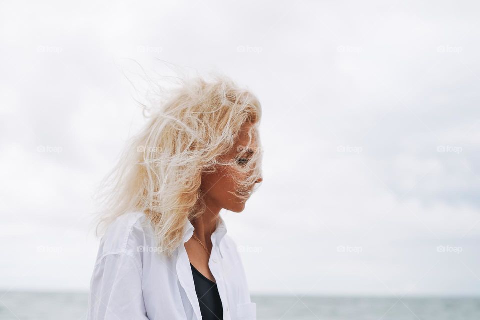 Portrait of Elegant blonde woman in white shirt on sand beach at storm sea at windy weater