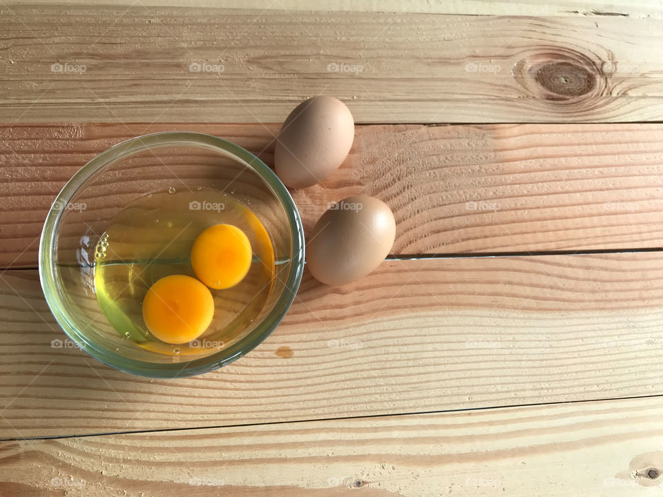 two fresh chicken yolk and egg white in glass cup and another two eggs beside it on natural rubber wood board with copy space on right side of frame