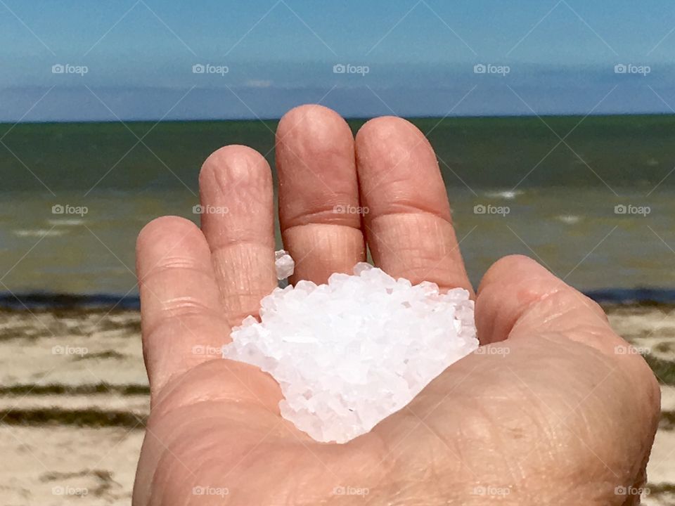 A closeup of a hand holding natural white rock salt in front of an ocean horizon on a clear day