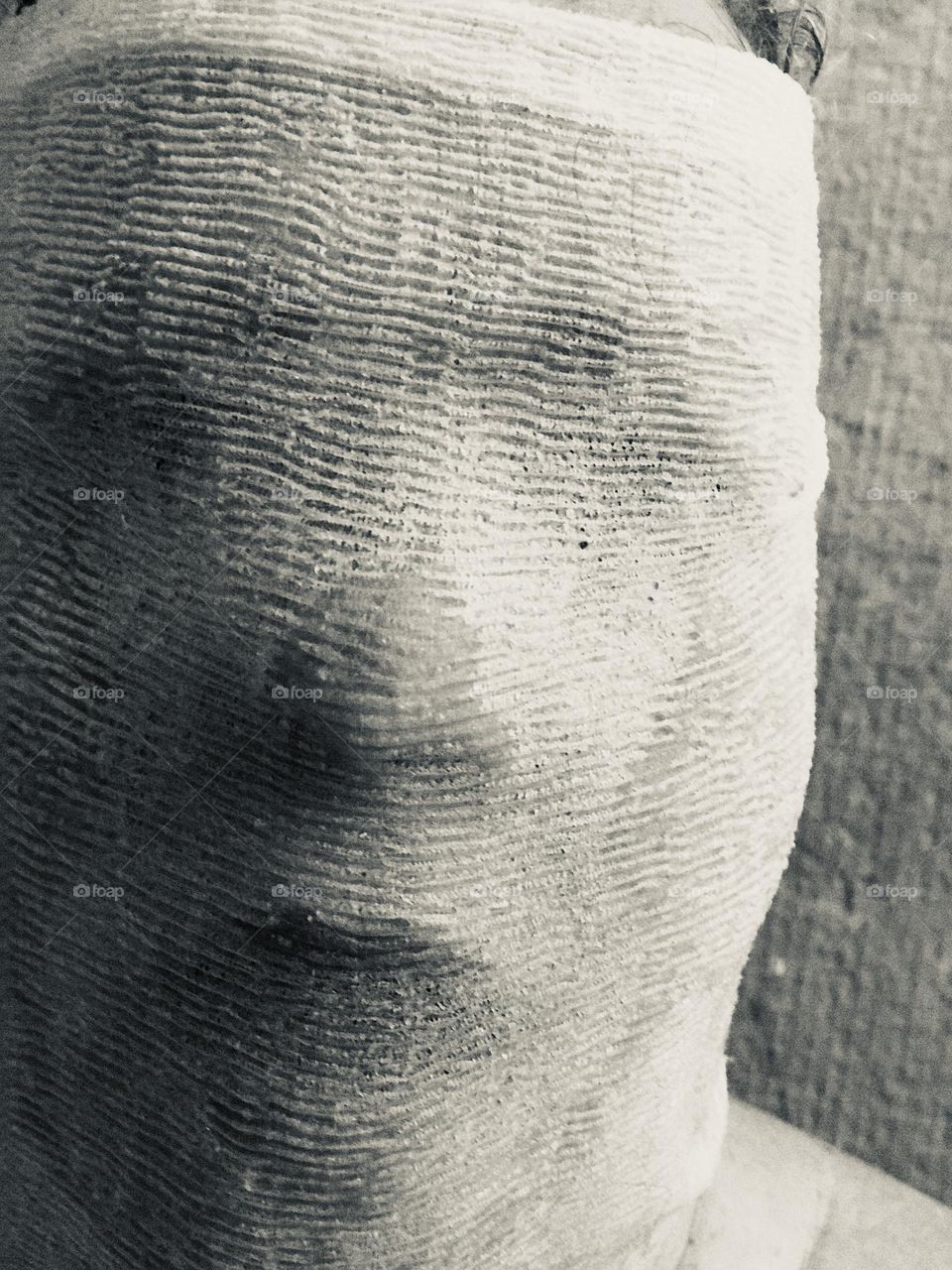 Black and white self-portrait of a woman’s head bound in a wet, textured like cheesecloth, mostly opaque, cloth, standing close to a textured wall. Her basic features are shown in highlight and shadow but she is indistinguishable. 