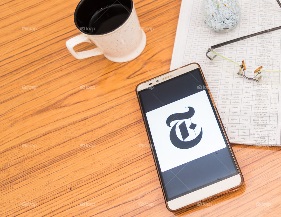 Kolkata, India, February 3, 2019: The New York Times news app visible on mobile phone screen beautifully placed over a wooden table with a newspaper and a cup of coffee. A Technology Product Shoot.