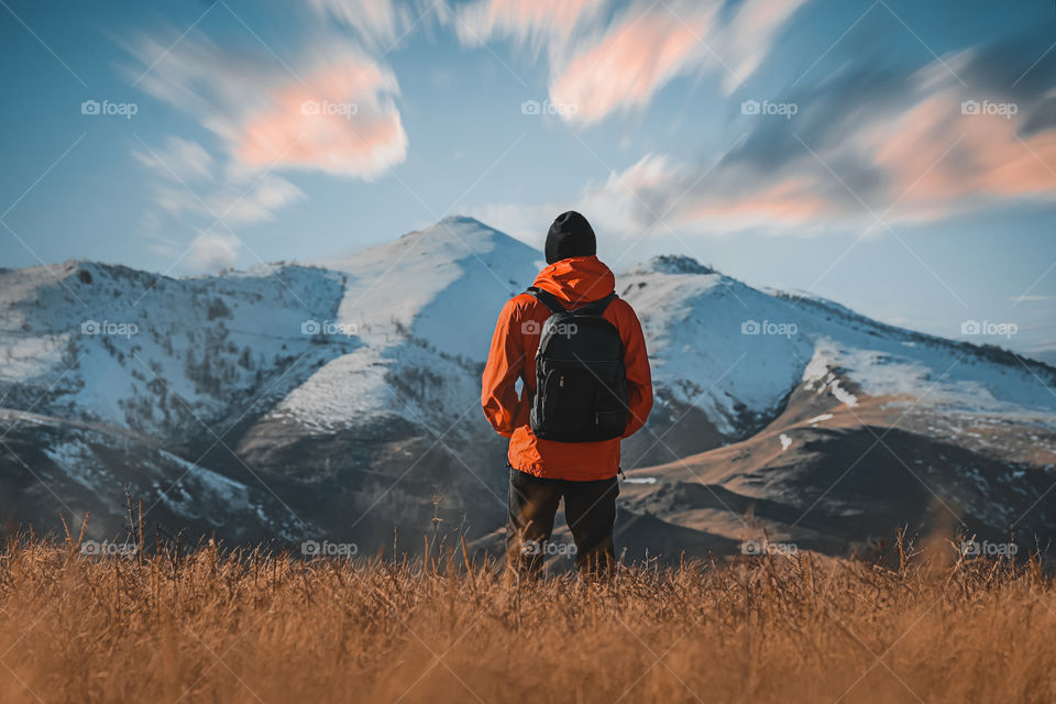 Rear view of male hiker standing on field against snowcapped mountains . 