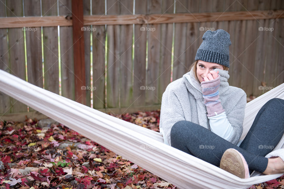 Young millennial woman sitting in a hammock outdoors in the fall wearing a beanie hat and fingerless gloves and playfully covering face