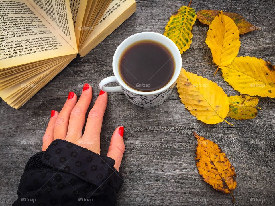 Cup of coffee on wooden table with yellow leaves and a book beside with a woman's hand with red nails