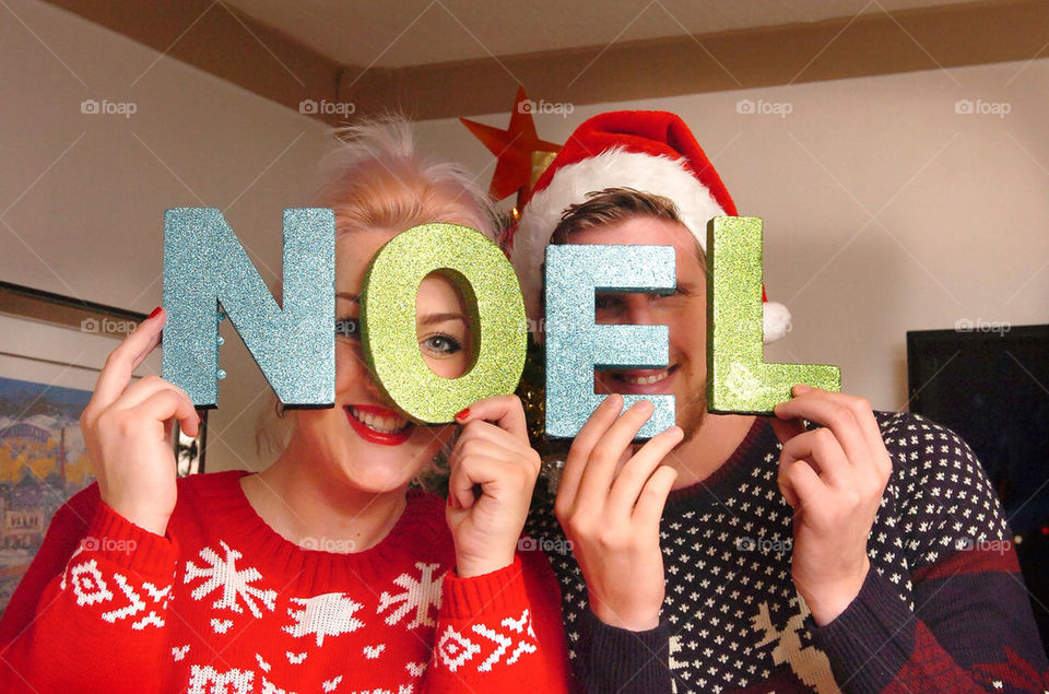 A young couple in Christmas knit ware hold up letters spelling NOËL