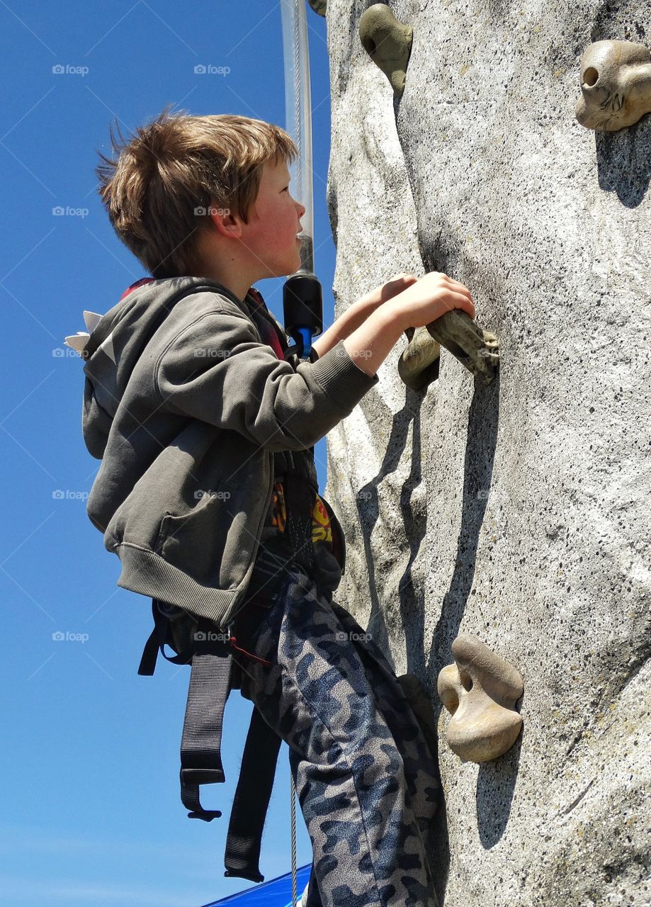 Boy On Rock Climbing Wall. Young Boy Using Harness And Ropes To Scale An Obstacle Wall
