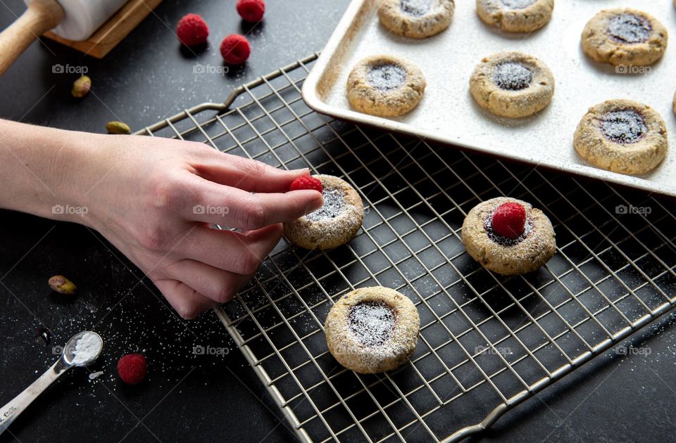 Person’s hand placing raspberries on freshly baked thumbprint cookies sprinkled with powdered sugar 