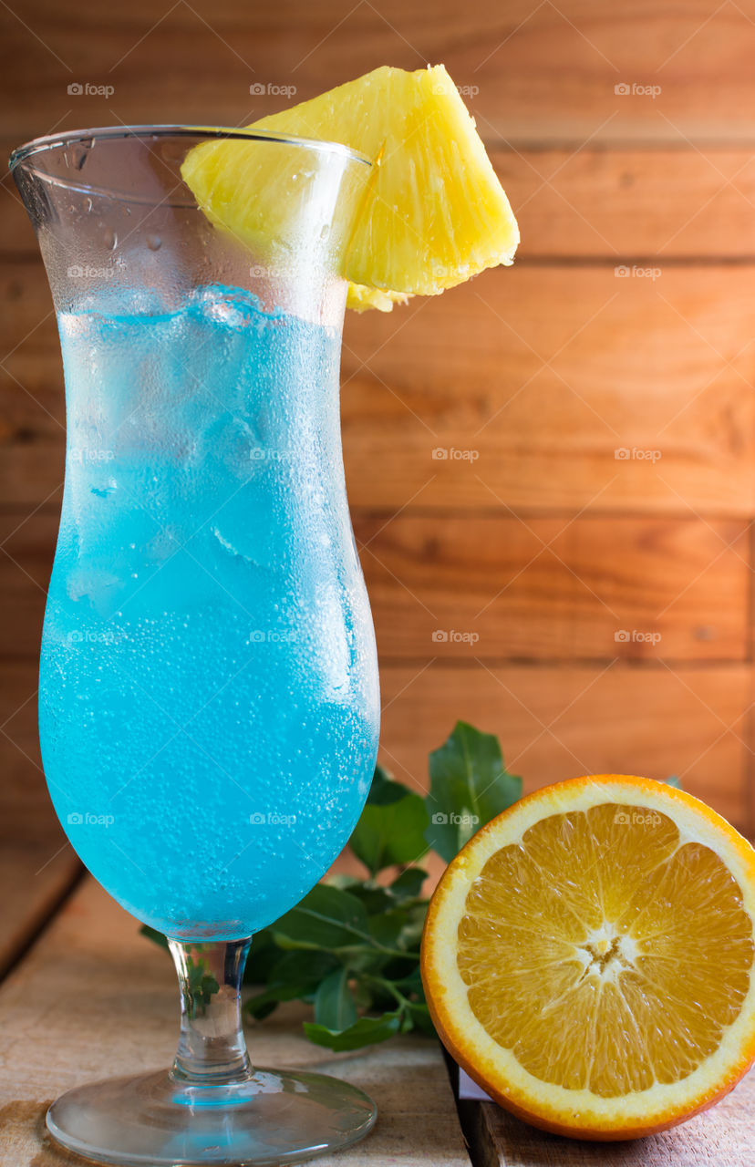 non-alcoholic fruity blue coctail served in an hour shaped glass with fresh pineapple on rim of glass and placed on a wood table with wood background and an orange sliced open next to the glass