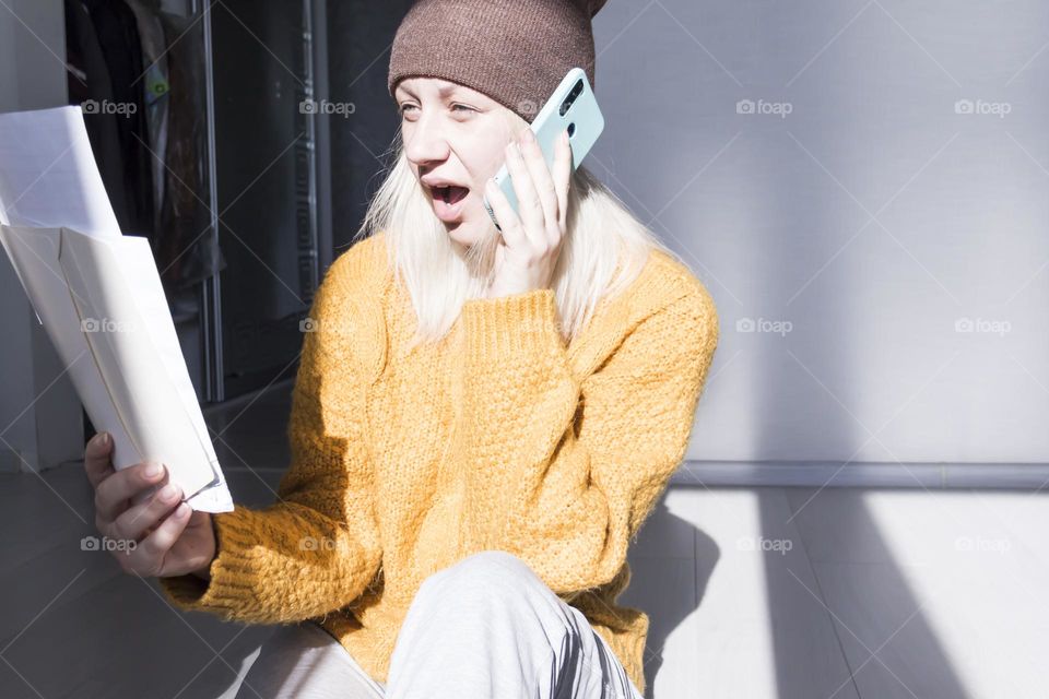 A young girl in a yellow sweater and hat is talking on a mobile phone and studying large bills and utility bills..