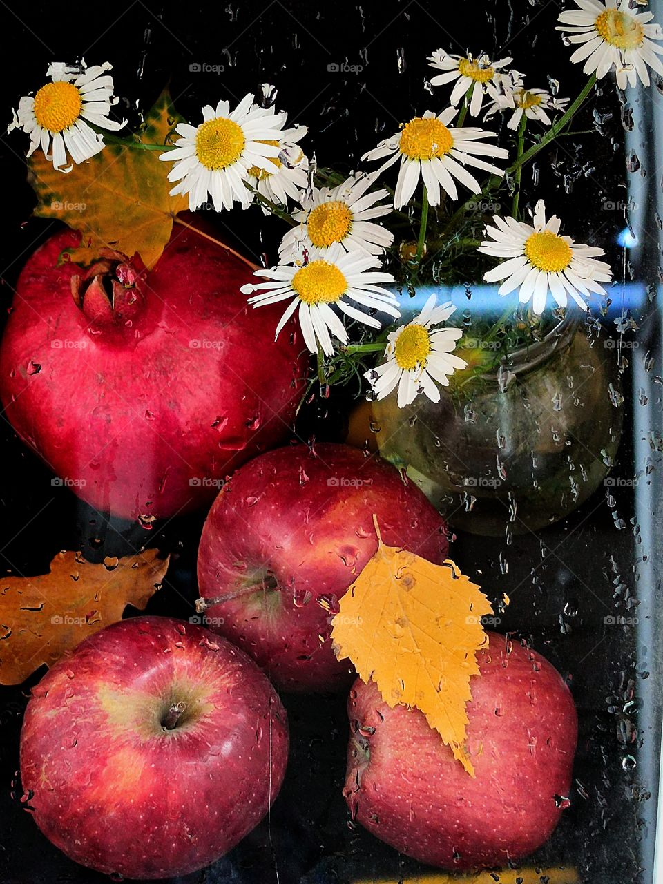 Autumn.  Window with raindrops.  Outside the window, a red pomegranate, red apples, a bouquet of daisies and yellow autumn leaves