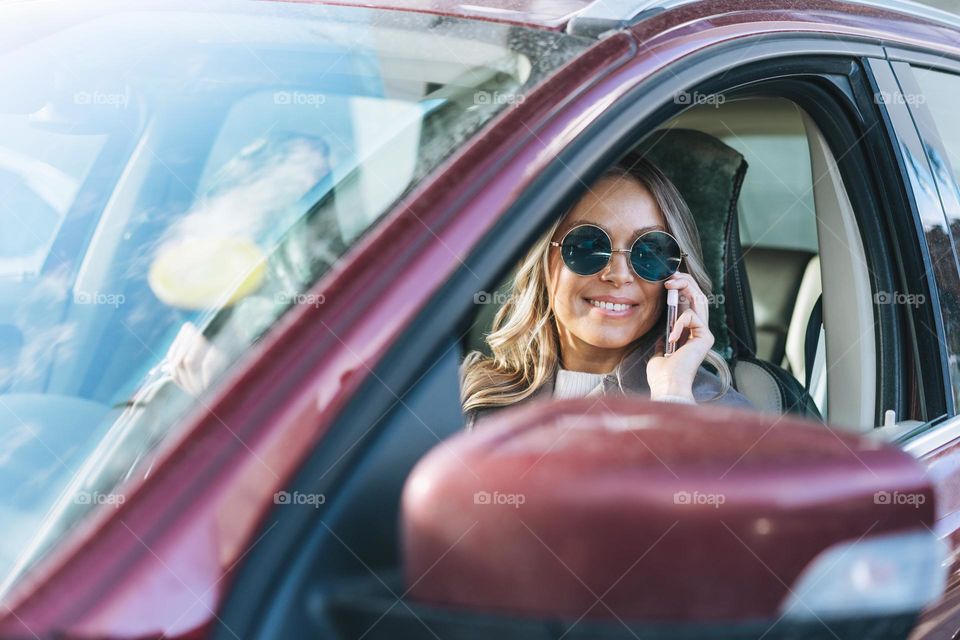 Young stylish woman with long blonde hair and sunglasses driving a car and using mobile phone