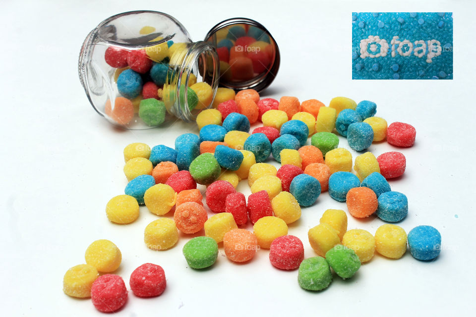 Sour chewy gummies flowing out of a small overturned glass jar. The up turned lid reflects the candies. A foap logo & foap name tag made of turquoise & white jelly beans is placed in the top right corner! ๐