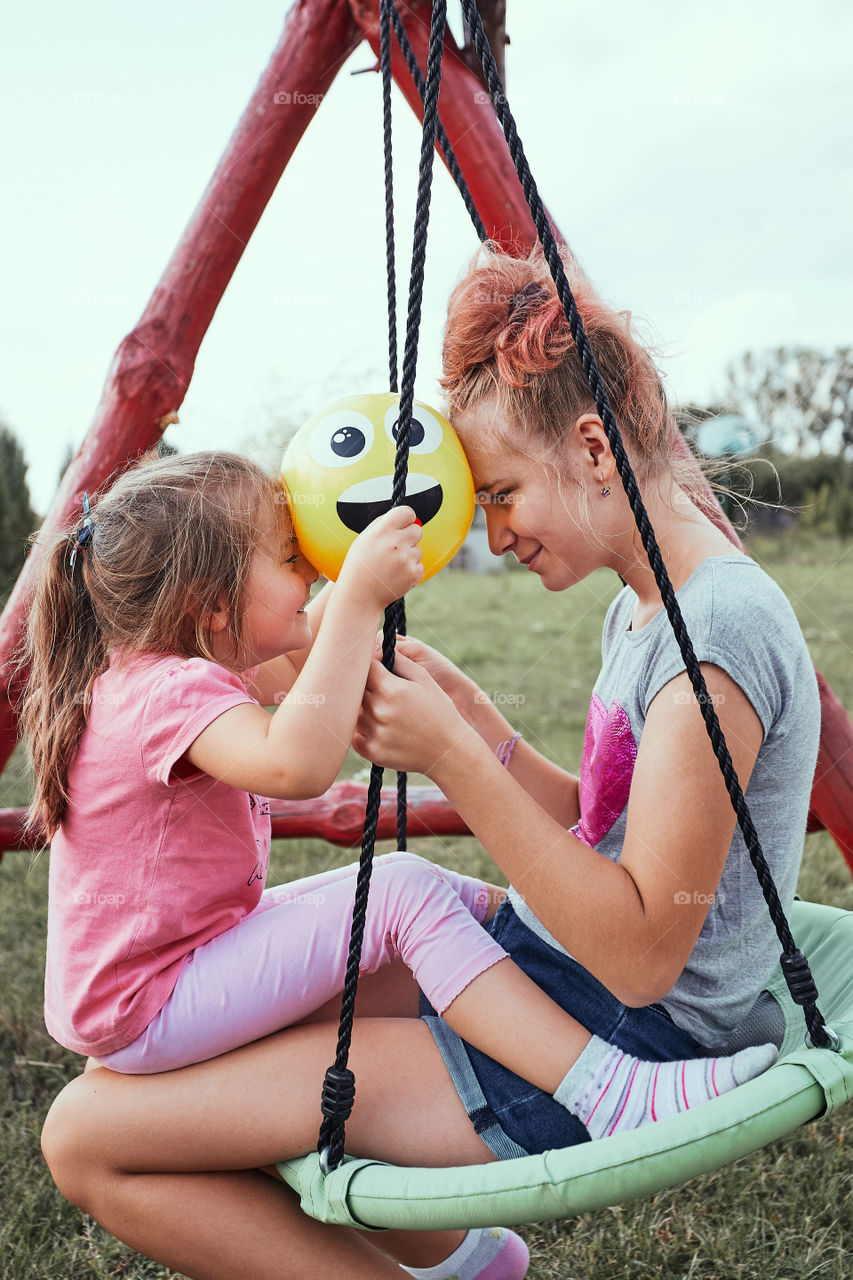 Teenage girl playing with her younger sister in a home playground in a backyard. Happy smiling sisters having fun on a swing together on summer day. Real people, authentic situations