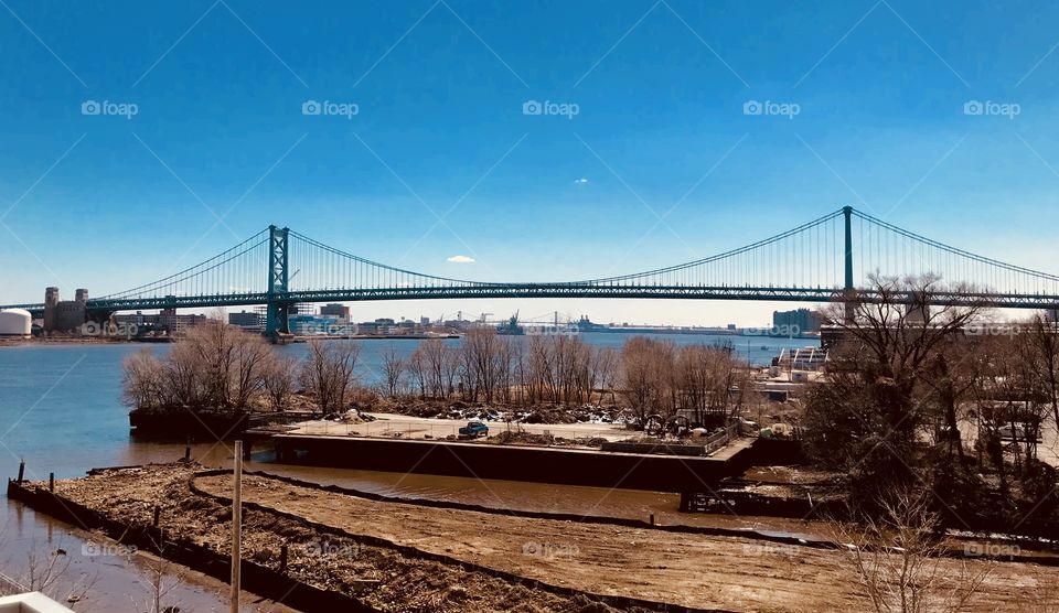 The Ben Franklin bridge from the Philadelphia side on the waterfront. The view from an apartment on penn ave. Bright blue sky over the water and bridge 