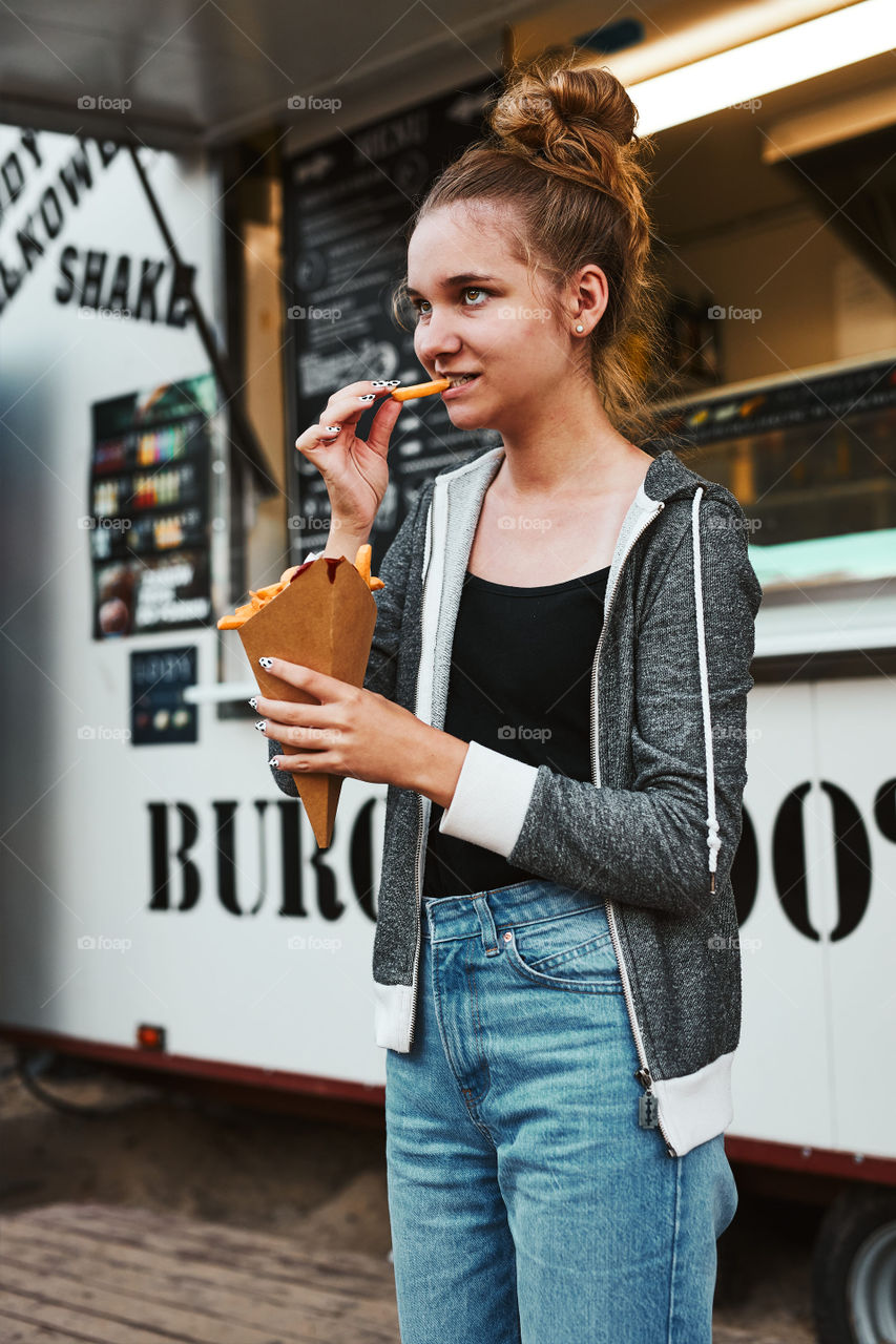 Teenage girl eating potato fries from carton cone standing in front of food truck. Teenager having a fast food meal outdoors during summer vacations