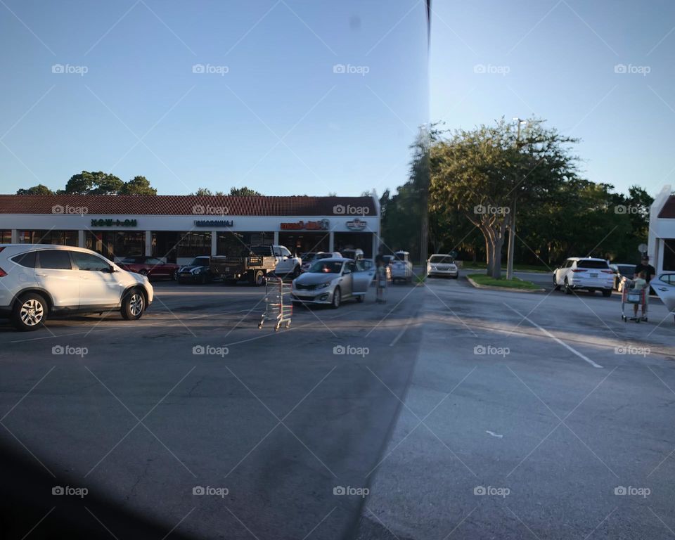 Commuting: many vehicles in a parking lot and a cart reflecting in a vehicle side mirror with a different perspective that seems to have two sides parking lot seeing businesses in the evening with sunset.