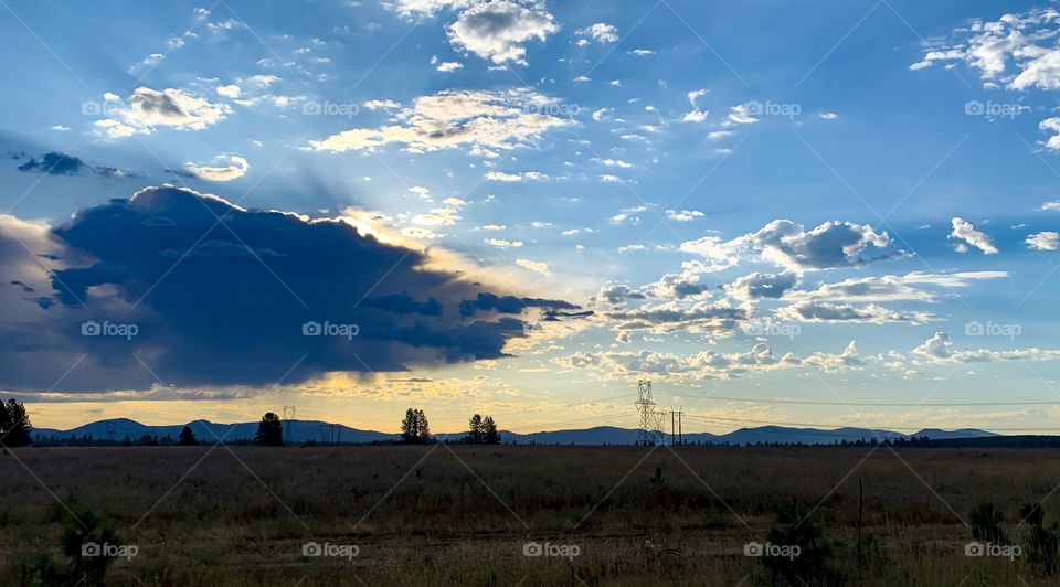 Sun rises behind a outward moving storm clouds. Yellow colors and sunbeams show from behind the dark cloud with bright blue sky and scattered clouds moving in. See the power towers and lines in the background, along with the mountain silhouette.