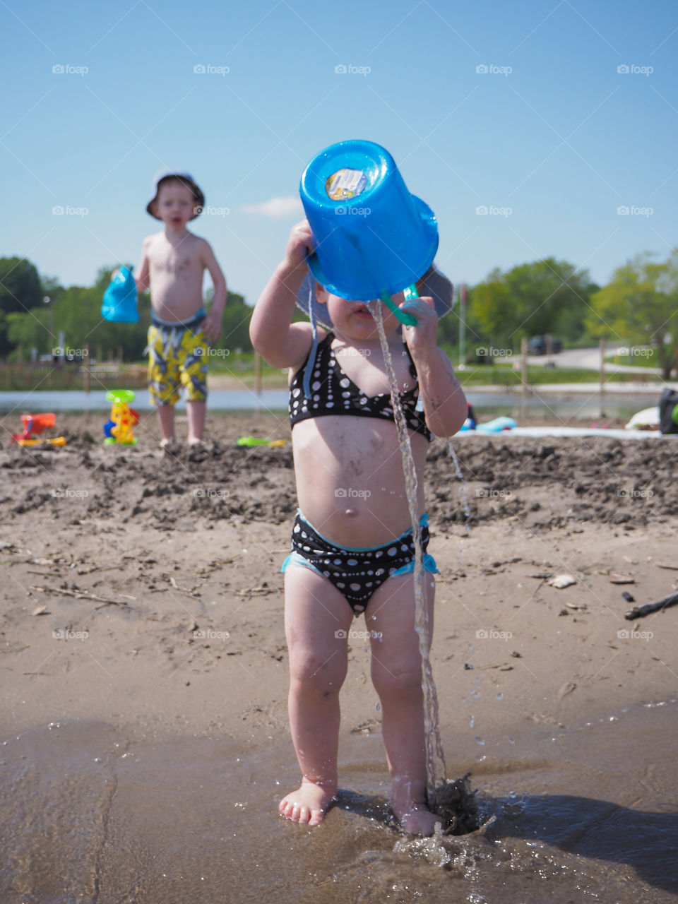 Toddler girl pours water on herself from bucket at the beach while brother watches in awe.