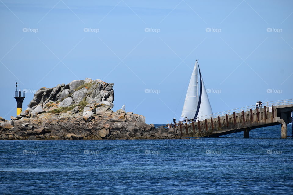 A ship with a sail near  sea and a bridge 