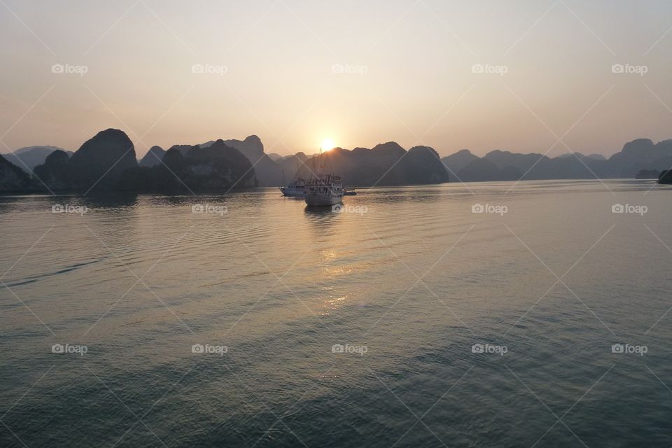 On the water there are 2 yachts , the water is slightly rippling.  Surrounding there are many undulating mountains, the sun is falling,shines into the water,creating beautiful light at sunset