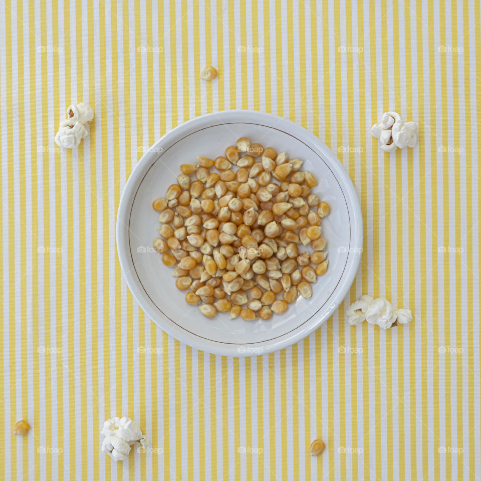 View from above white dish with popcorn grains in background with white and yellow striped pattern and with some white popcorn