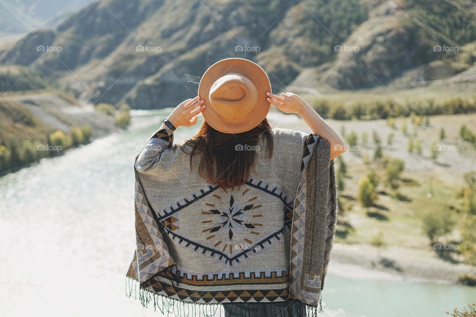 Carefree brunette young woman traveler in poncho from back on background of mountain river