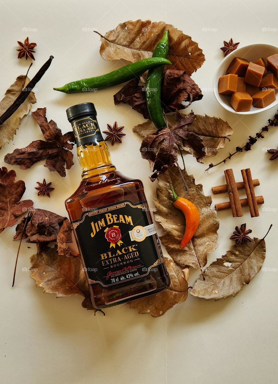 Jim Beam Black Extra Aged bourbon product flatlay on cream surface and autumn leaves with fresh peppers, cinnamon sticks, vanilla pod and caramels in a china bone bowl