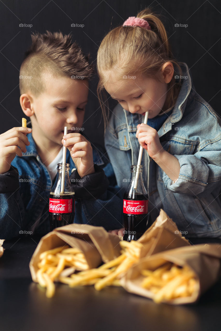 stylish children, dressed in denim clothes, drinking Coca-Cola. They eat, relax, communicate at home. sister and brother drink coca cola