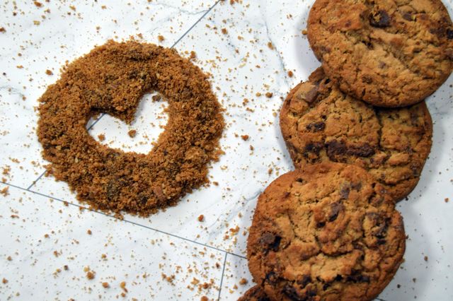 A simple heart hollowed out of the centre of a sprinkling of cookie crumbs sits beside a pile of chocolate chip cookies.
