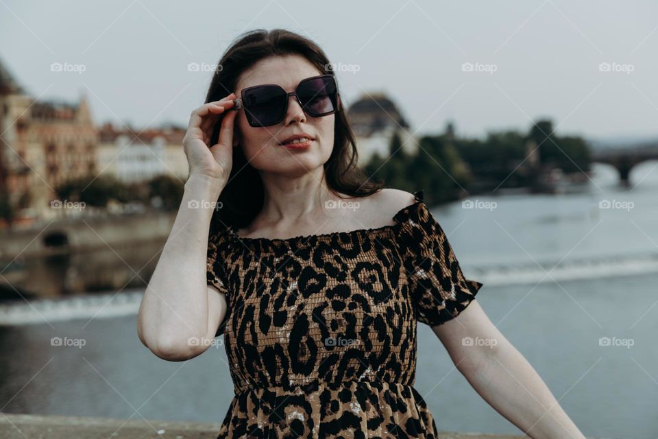 Portrait of one beautiful Caucasian brunette girl in tiger print dress and sunglasses looking away with smile standing on Charles Bridge in Prague on summer sunset evening, side view close up.