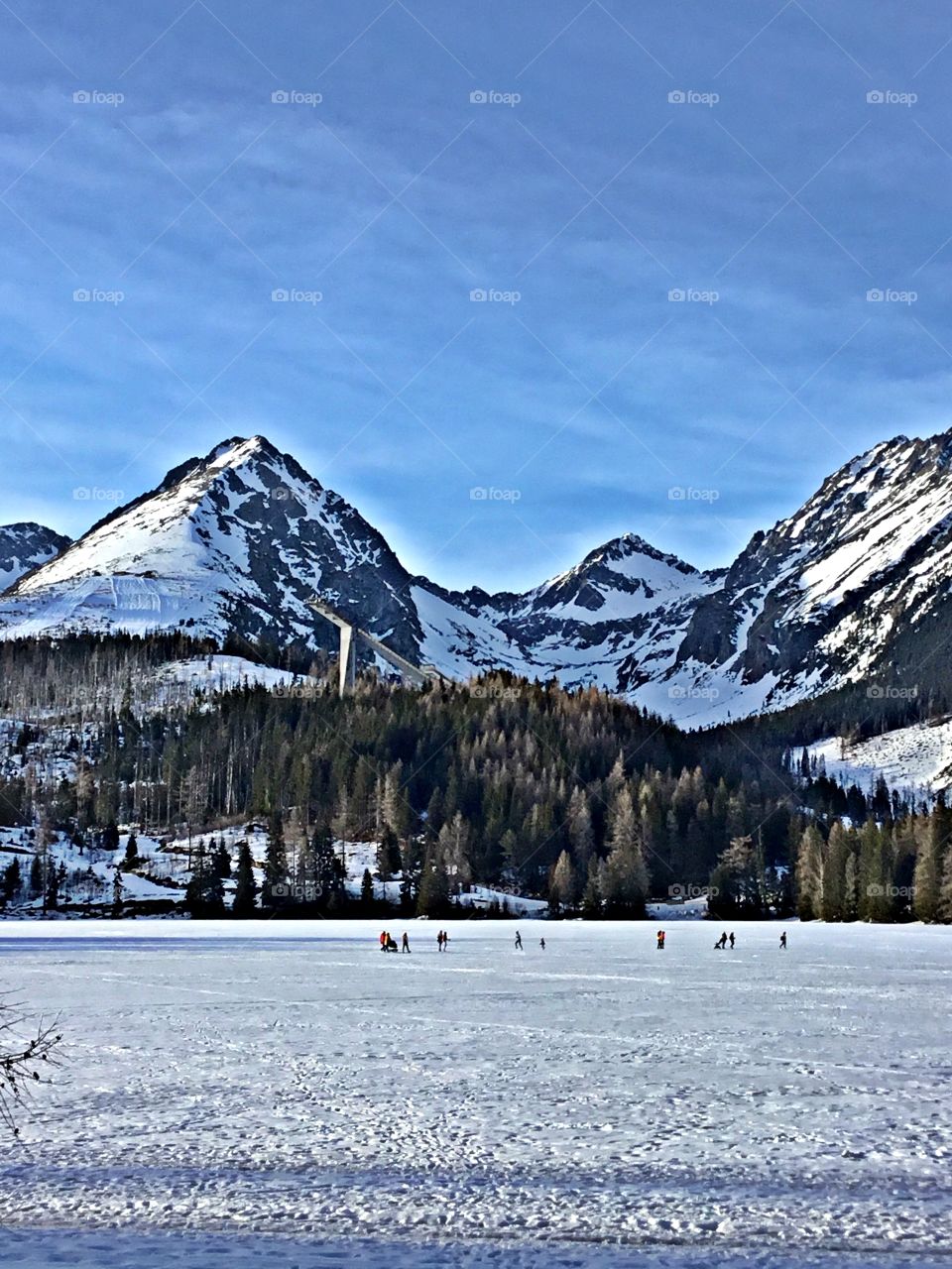 Landscapes of 2019 - Foap Missions - A view of people walking across a frozen lake at the base of the snow capped High-Tatry Mountains 