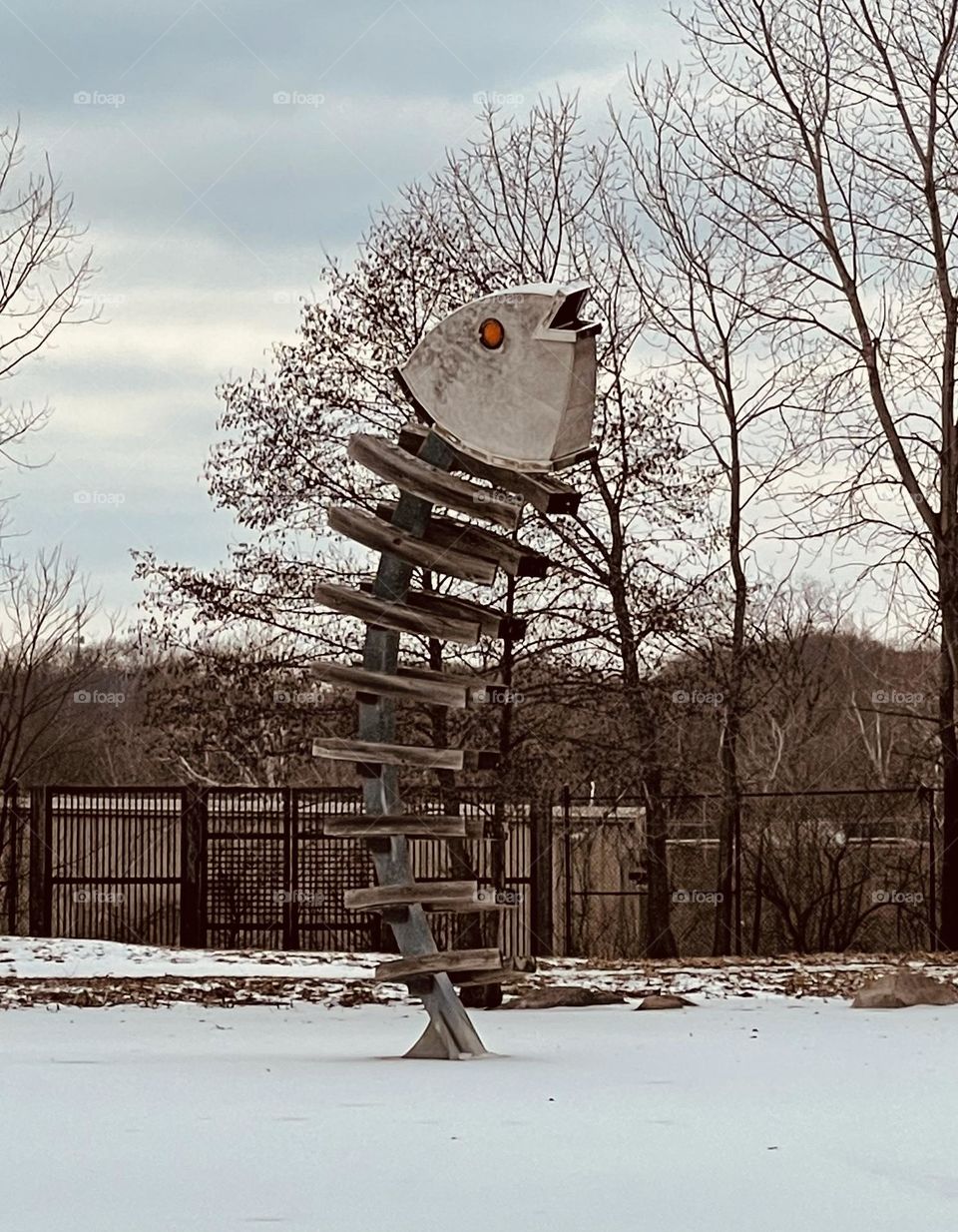 A gigantic wood and steel sculpture of a fish, at the back of a small frozen pond. 