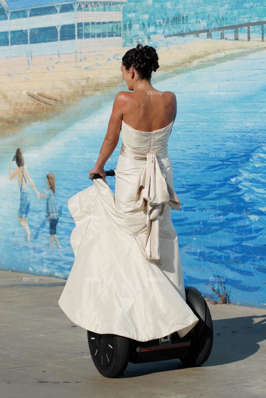 A bride in off white riding a Segway on a sunny day against a light blue painted wall