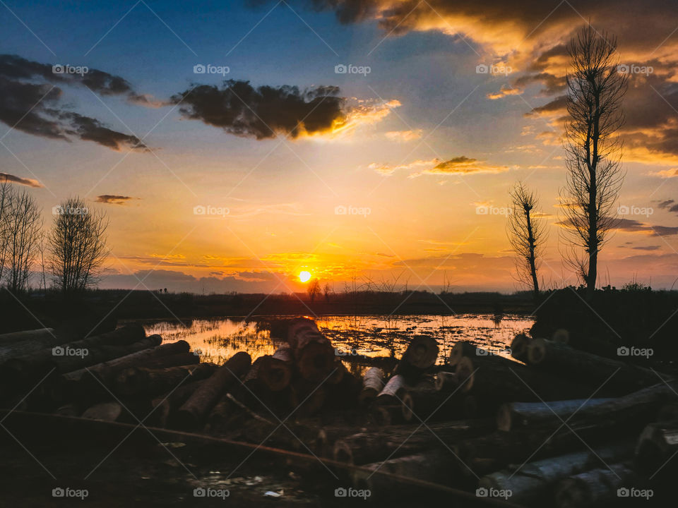 Stunning sunset near the lake. The sky is partly cloudy. The wood logs are visible by the lake.