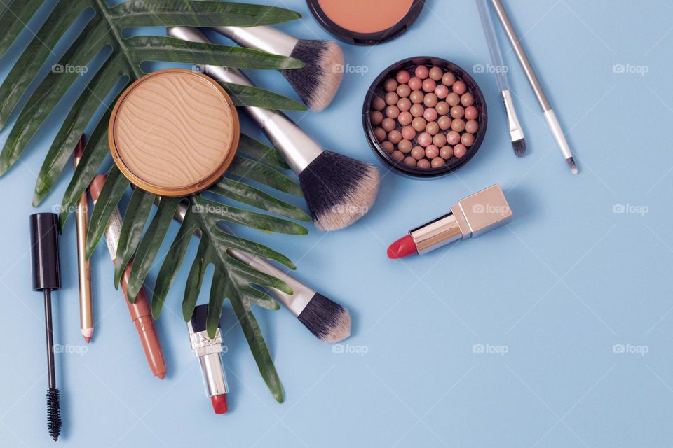 Makeup set of pallet of nude eyeshadow, round shaped face powder, red lipstick, soft black eyebrow shadow, makeup brushes and eyebrow pencil with palm tree branch on blue background, flat layered close-up. Concept female cosmetics, beauty salon.