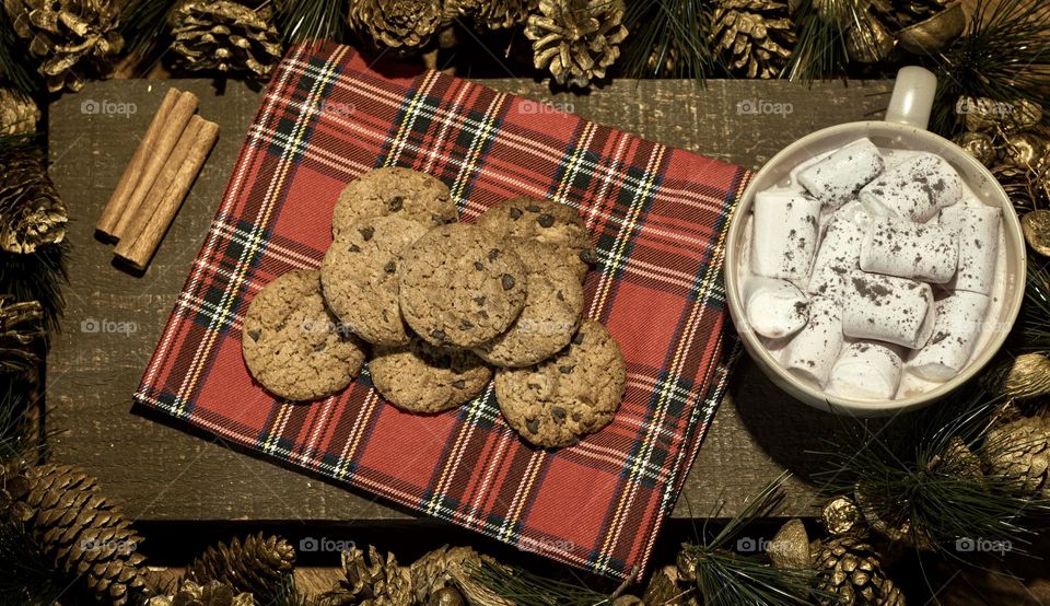 Christmas cookies on a tartan napkin with hot chocolate and marshmallows, presented on a wooden board surrounded by gold pine cones.