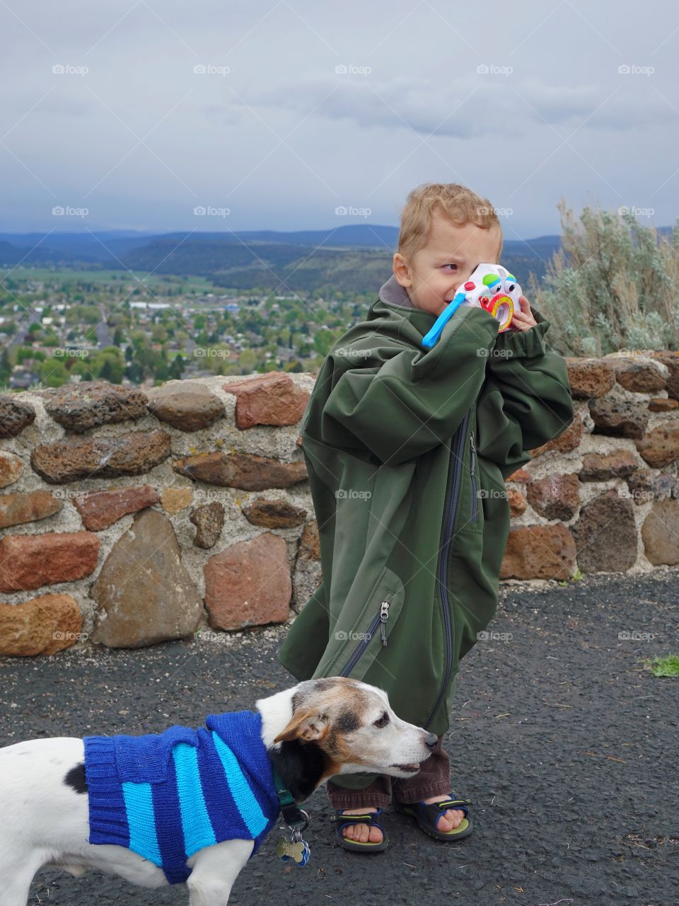 A cute young boy in an adult jacket that hangs to the ground along with a Jack Russell Terrier in a blue sweater takes a photo with his toy camera. 